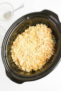 A slow cooker filled with a casserole topped with crushed crackers or bread crumbs, next to an empty glass bowl with a spoon.