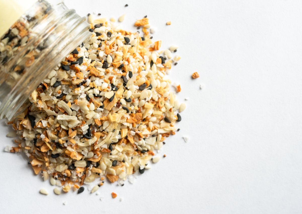 A jar tipped over, spilling a mix of sesame seeds, dried garlic, dried onion, and poppy seeds onto a white surface.