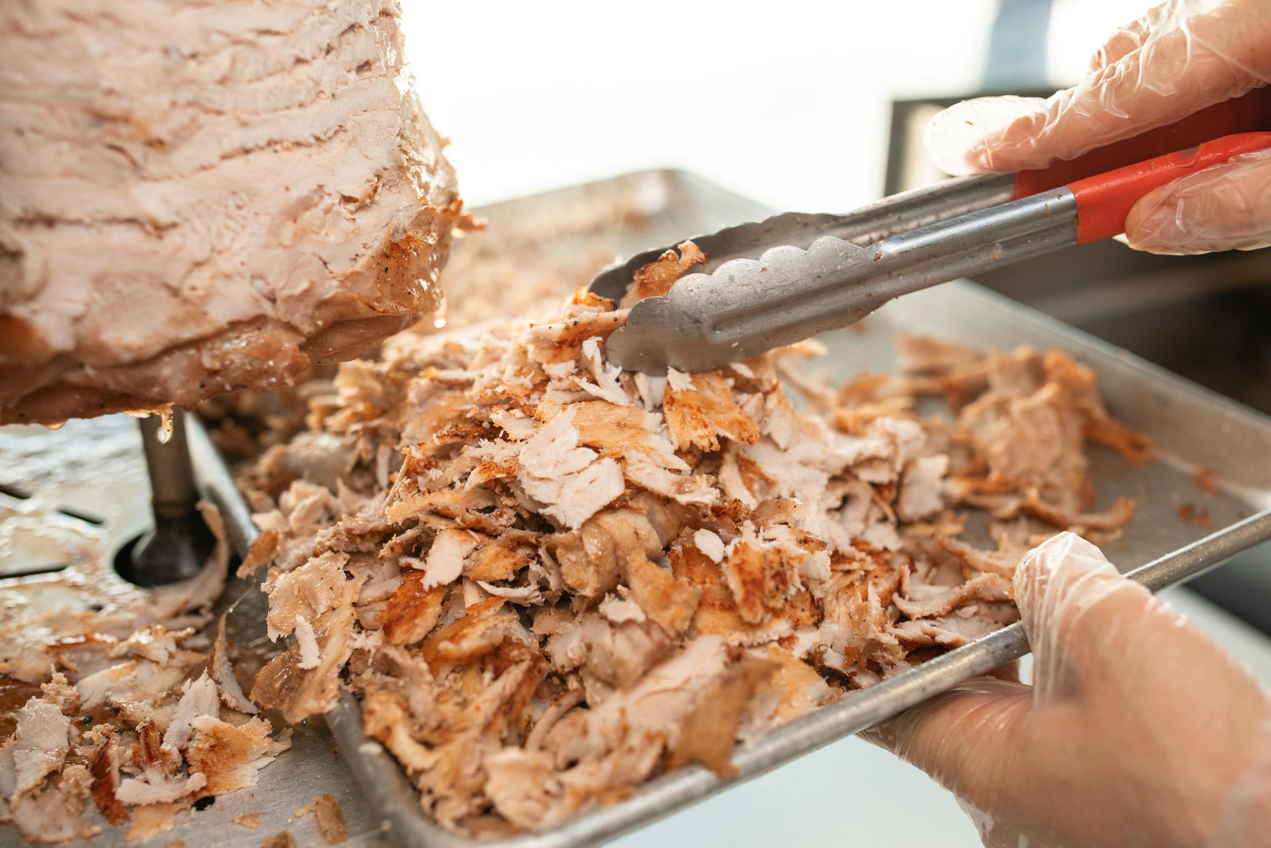 Freshly sliced shawarma meat on a tray being prepared with tongs in a kitchen setting.