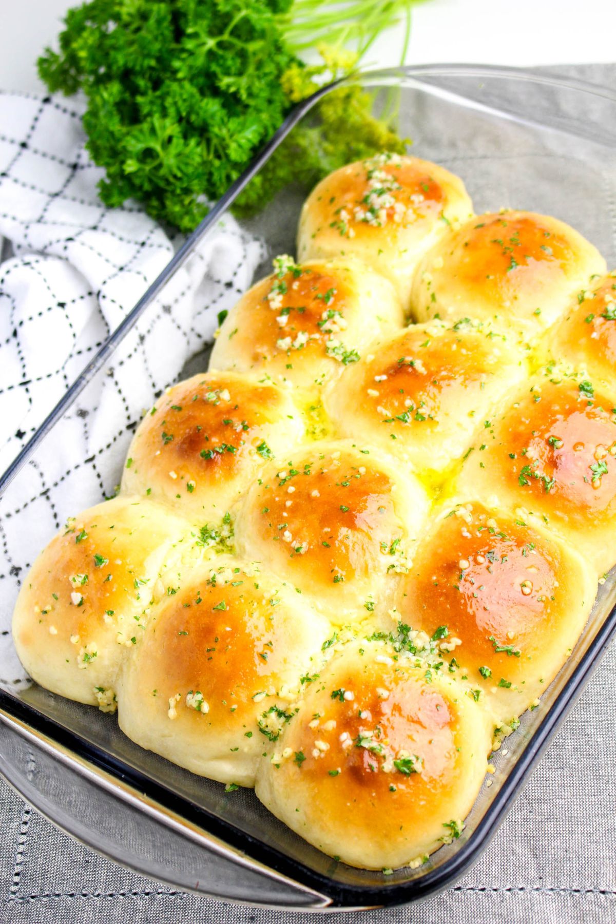 A glass baking dish filled with twelve golden brown garlic rolls topped with chopped herbs and garlic, with fresh parsley in the background.