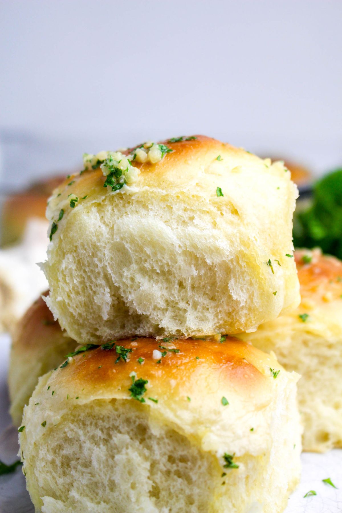 Two golden brown, fluffy dinner rolls stacked, topped with chopped parsley and garlic, with a blurred background.
