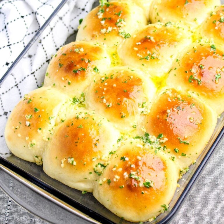 A glass baking dish filled with golden-brown garlic rolls topped with chopped herbs and melted butter, resting on a patterned cloth.