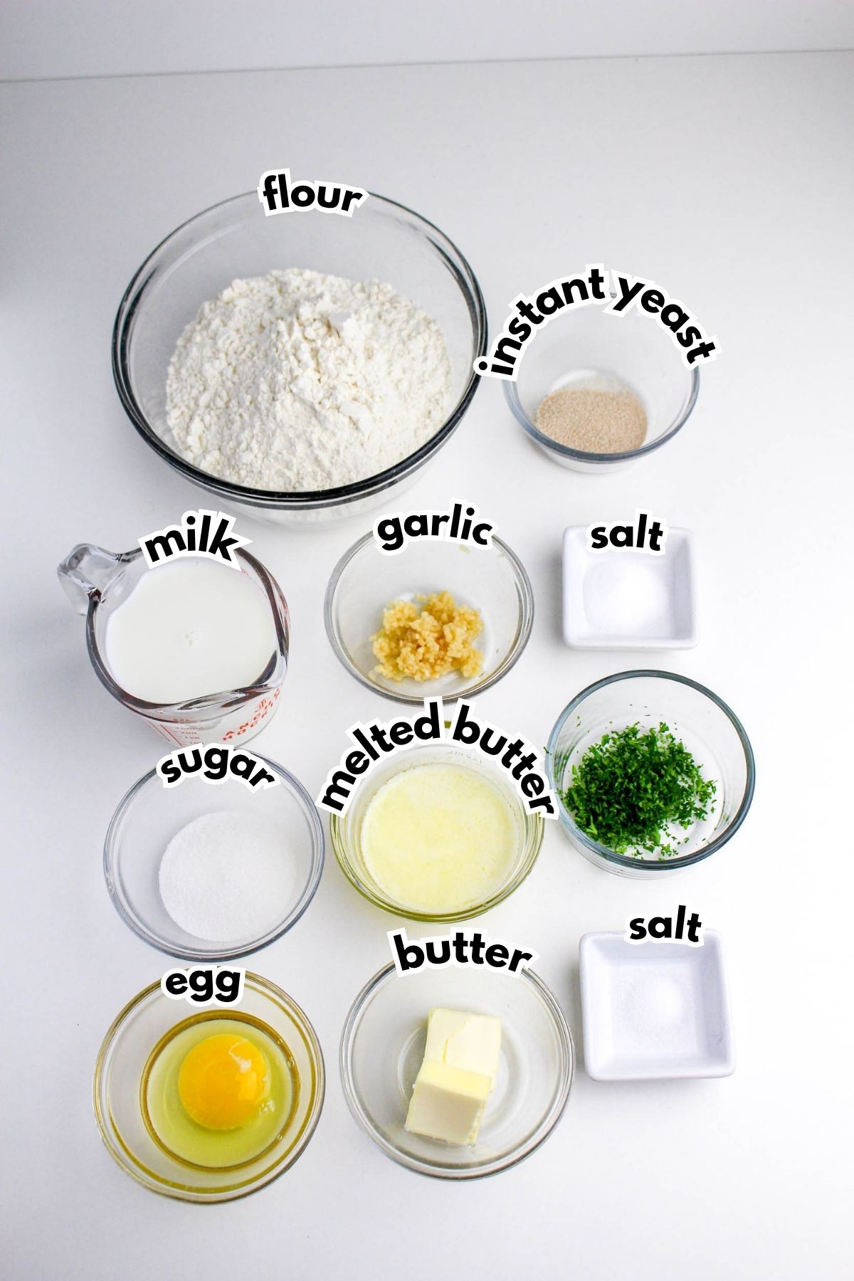 A collection of labeled ingredients in bowls and cups including flour, instant yeast, milk, garlic, salt, sugar, melted butter, parsley, butter, and an egg, arranged on a white surface.