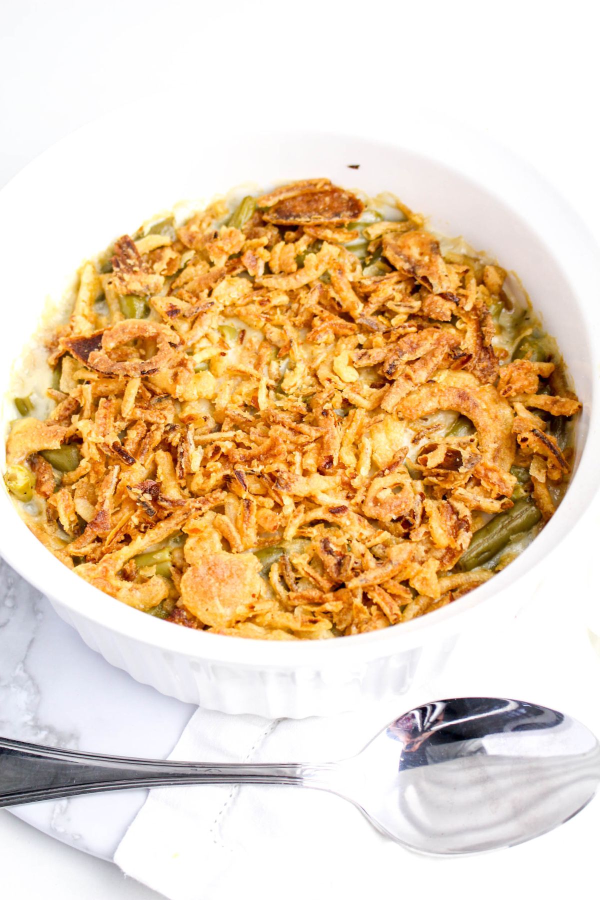 A white casserole dish filled with green bean casserole topped with crispy fried onions, placed next to a metal serving spoon on a white surface.