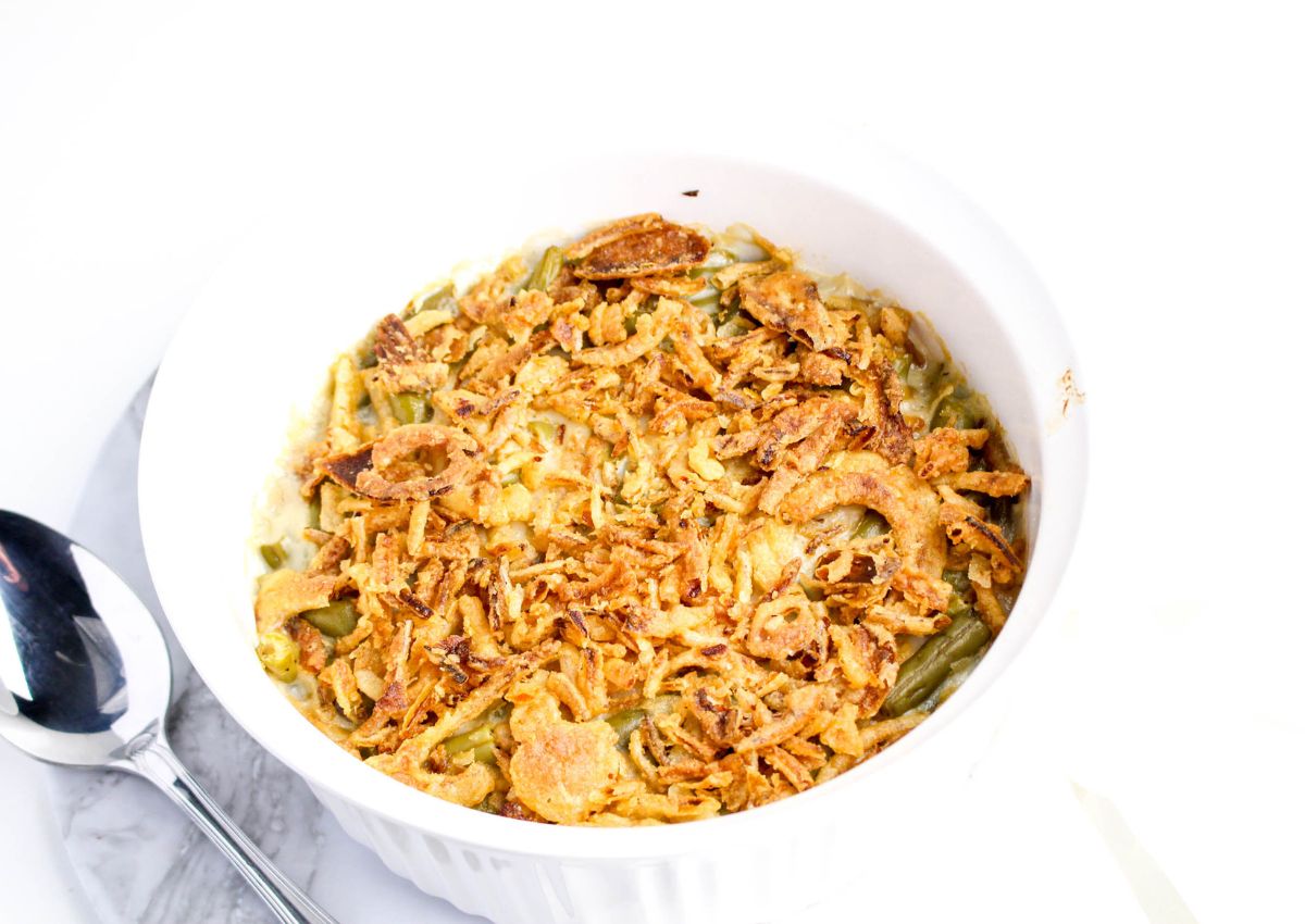 A white baking dish filled with green bean casserole topped with crispy fried onions, next to a spoon on a marble surface.
