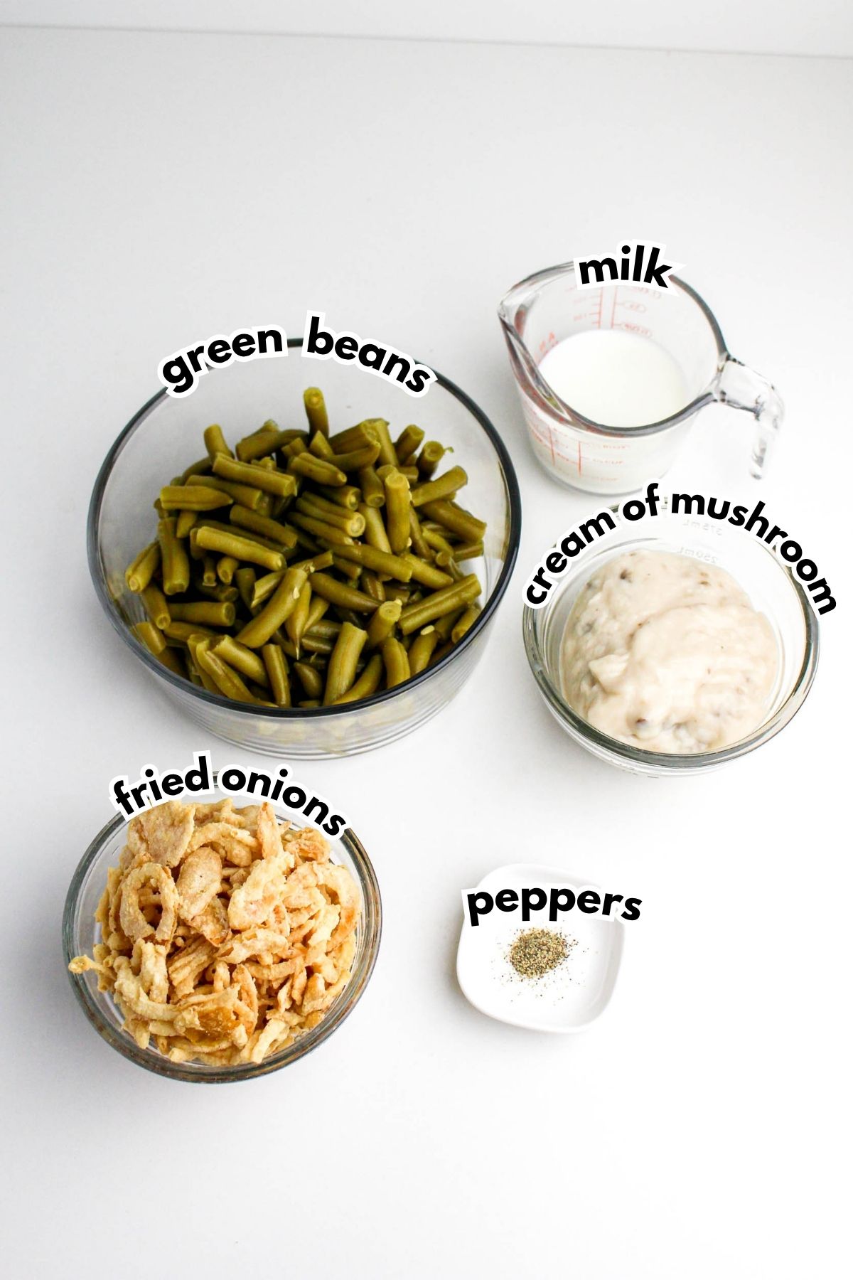 Four labeled bowls on a white surface containing green beans, fried onions, cream of mushroom soup, milk in a measuring cup, and a small dish of peppers.