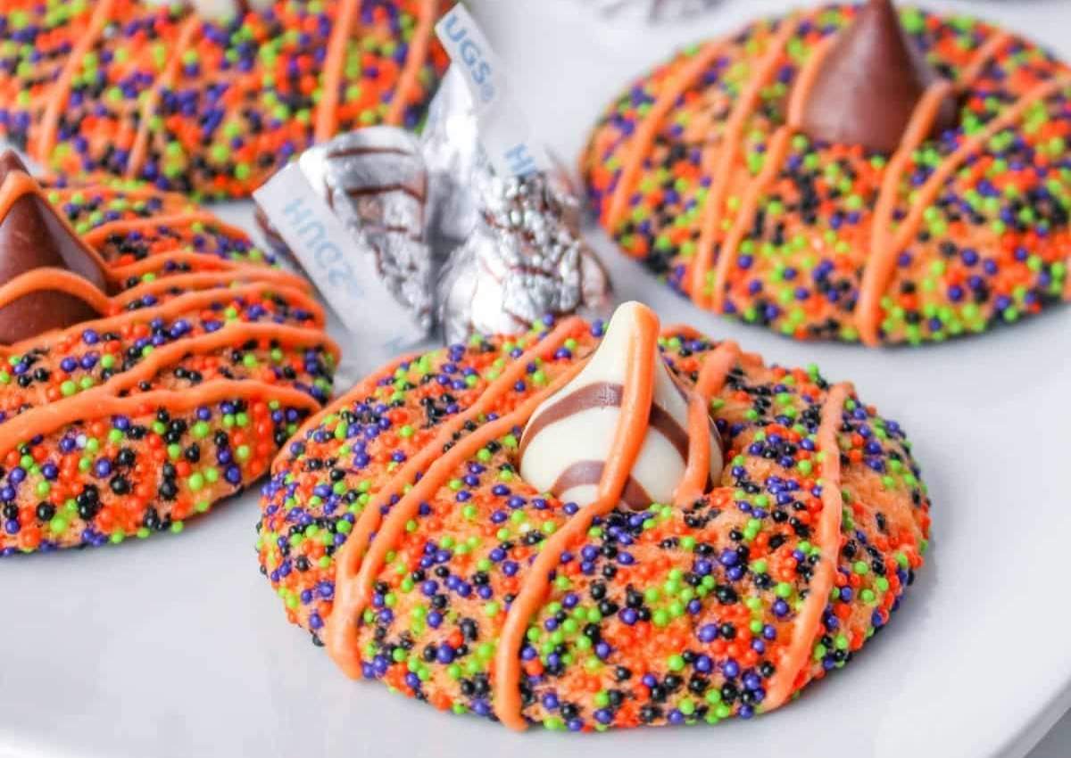 Colorful cookies covered in orange, green, purple, and black sprinkles, topped with chocolate kisses, and drizzled with orange icing on a white plate.