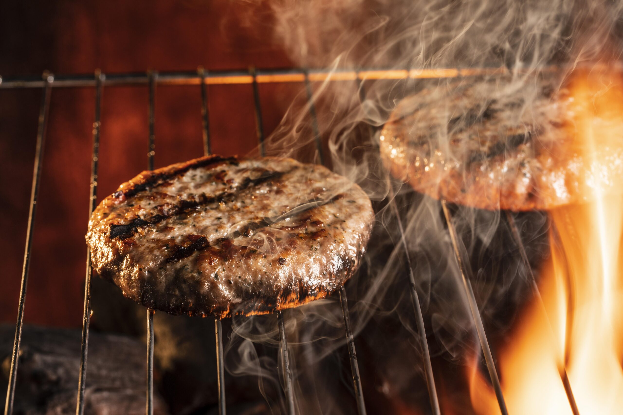 Two beef burger patties cooking on a grill with visible flames and smoke rising around them.