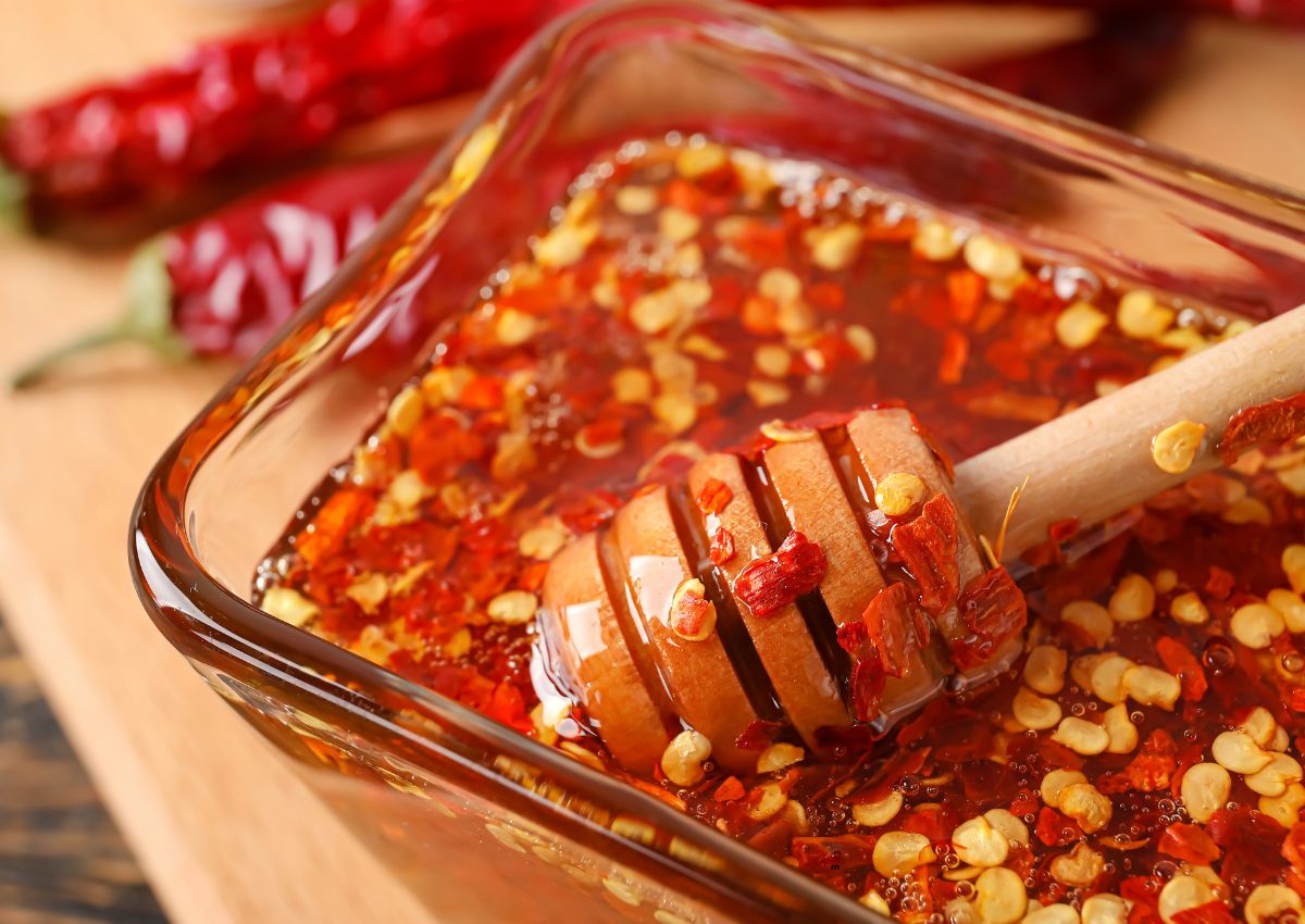 A glass dish filled with chili-infused honey, mixed with red chili flakes, and a wooden honey dipper partially submerged.