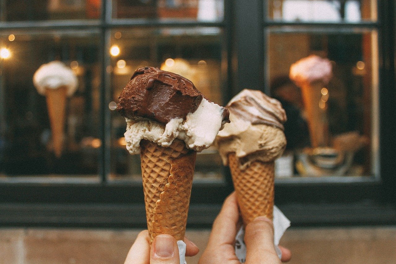 Two hands holding ice cream cones with multiple scoops, in front of a shop window displaying cones in the background.