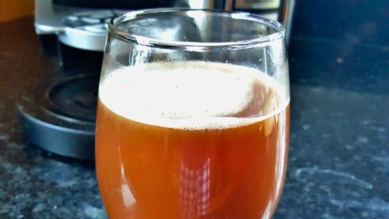 A glass filled with a brown, frothy liquid is placed on a dark countertop near a kitchen appliance.