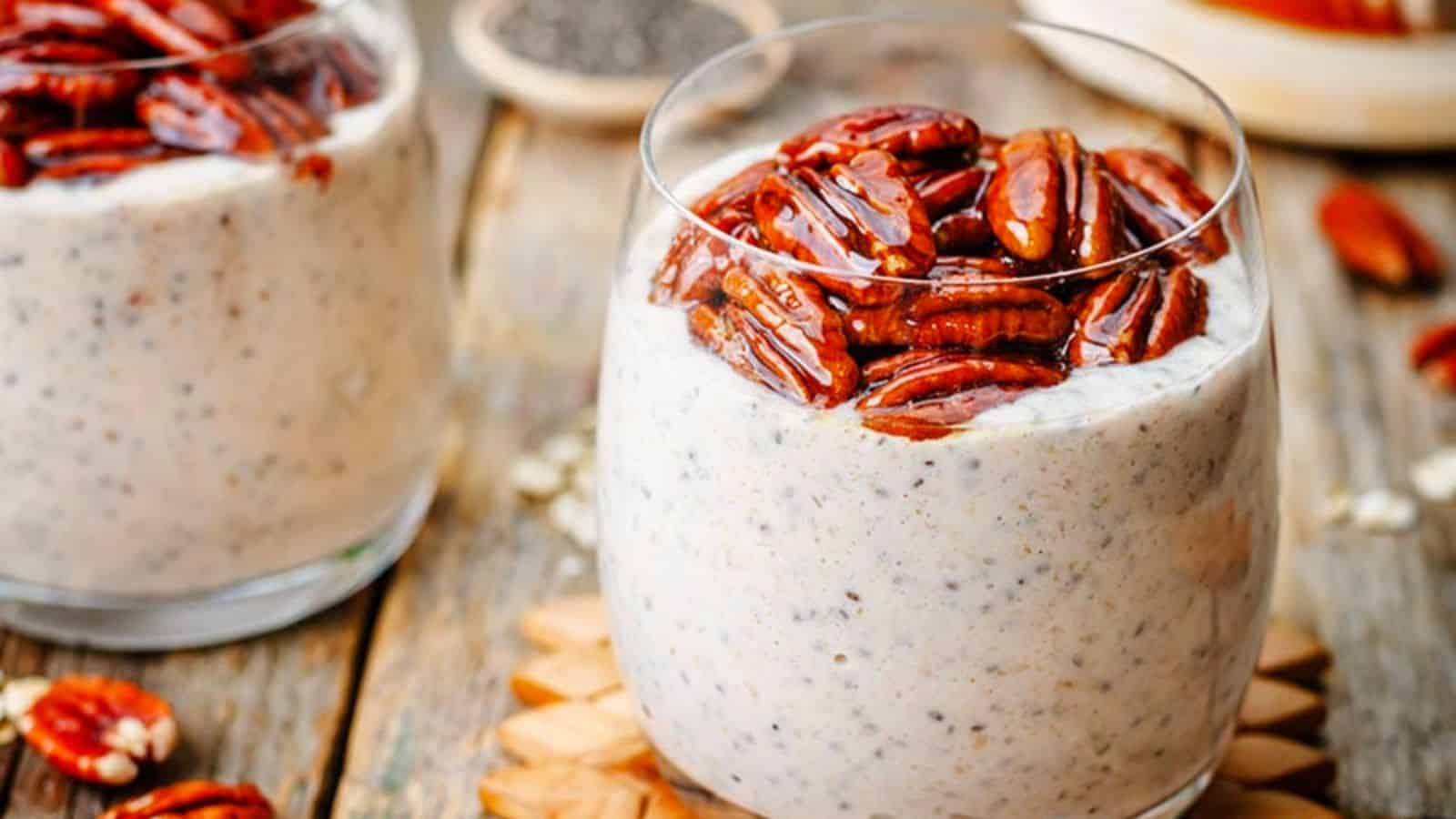 A glass filled with chia pudding topped with glazed pecans, placed on a wooden coaster with another glass and ingredients in the background.