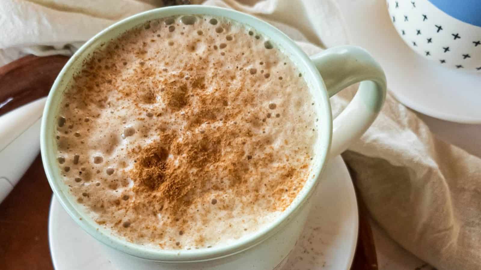 A close-up of a mug filled with frothy coffee topped with cinnamon, placed on a white saucer.