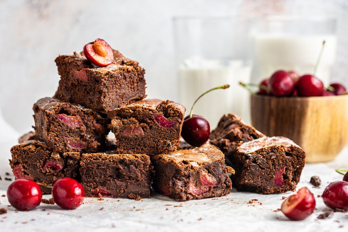 A stack of chocolate brownies with cherries, surrounded by fresh cherries, a bowl of cherries, and two glasses of milk in the background.