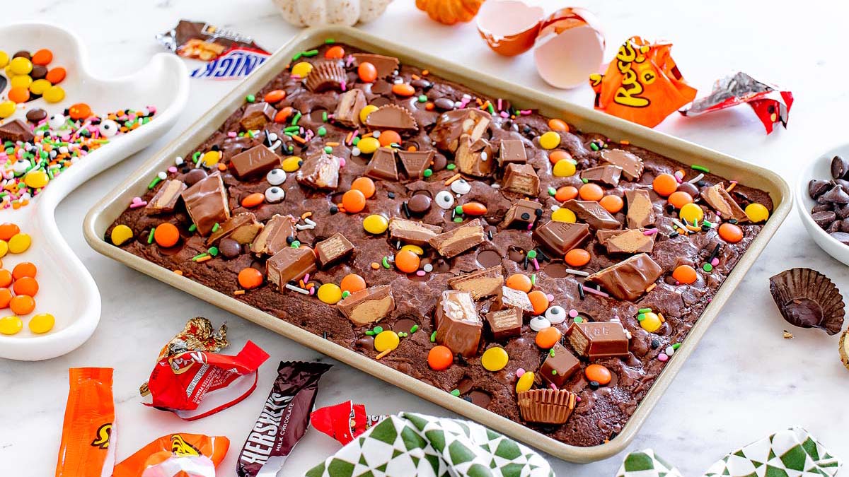 A baking tray of chocolate brownies topped with assorted Halloween candies, candy eyes, and sprinkles, surrounded by candy wrappers and baking ingredients on a white surface.