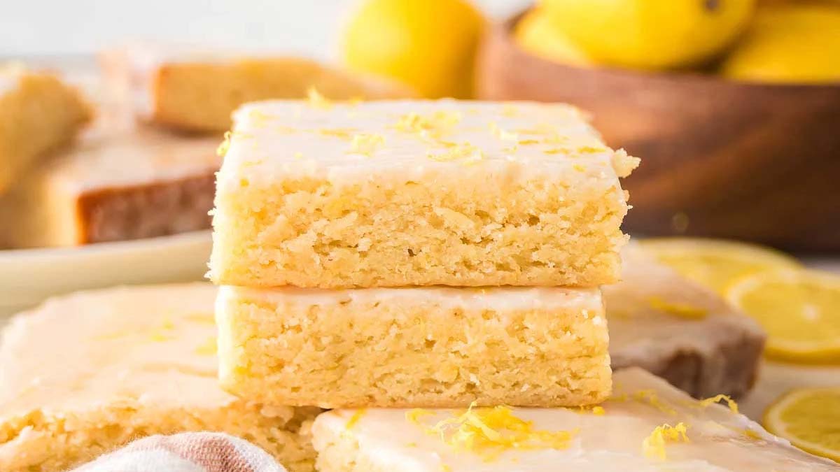 Two stacked lemon bars with icing and lemon zest on top, with more lemon bars and whole lemons in the background.