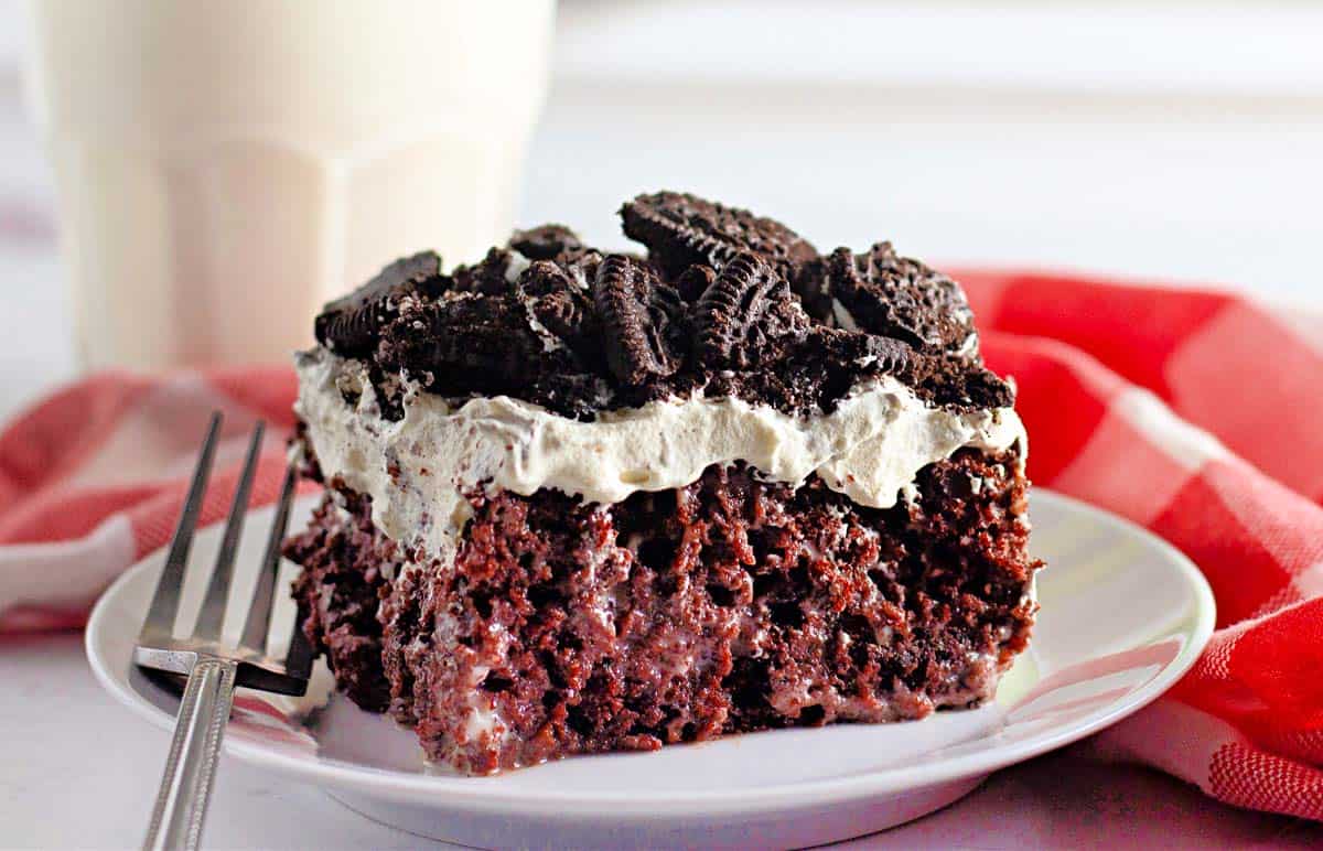 A slice of chocolate poke cake topped with whipped cream and crushed chocolate cookies on a white plate with a fork, next to a red and white napkin.