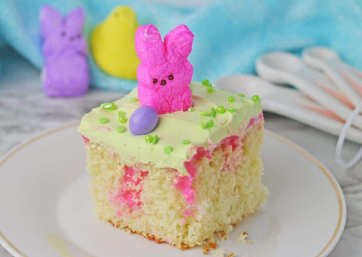 A square piece of vanilla cake with green frosting, a pink marshmallow bunny, green sprinkles, and a purple candy on top. Purple and yellow marshmallow chicks are in the background.