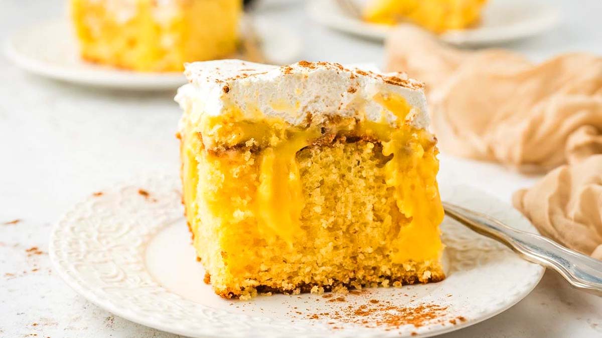 A slice of yellow poke cake with creamy filling and whipped topping sits on a white plate with a fork.