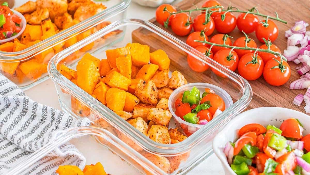 Glass meal prep containers with seasoned chicken, roasted sweet potatoes, and a small cup of tomato salad. Fresh cherry tomatoes and chopped vegetables on a cutting board nearby.