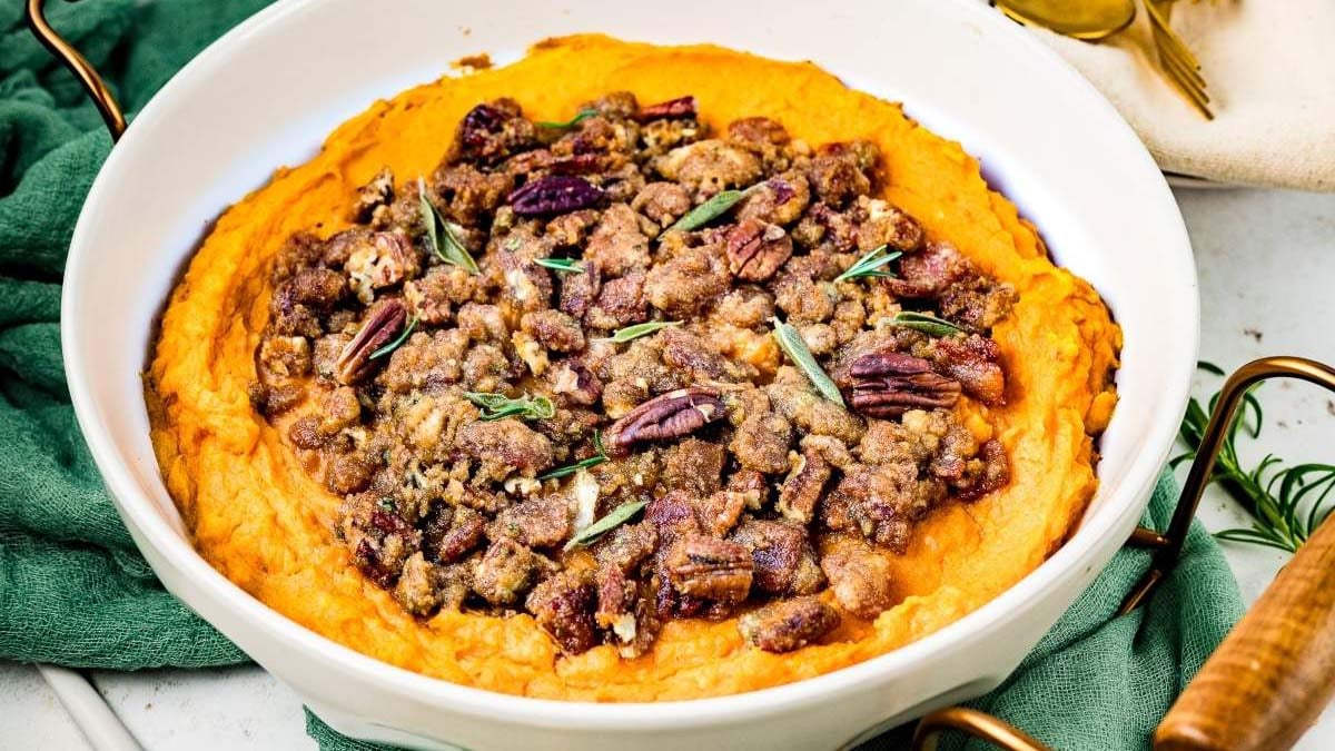 A white dish filled with mashed sweet potatoes topped with a pecan crumble and garnished with fresh herbs.