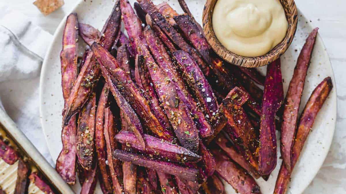 A plate of baked purple sweet potato fries seasoned with spices, served with a bowl of creamy dipping sauce.