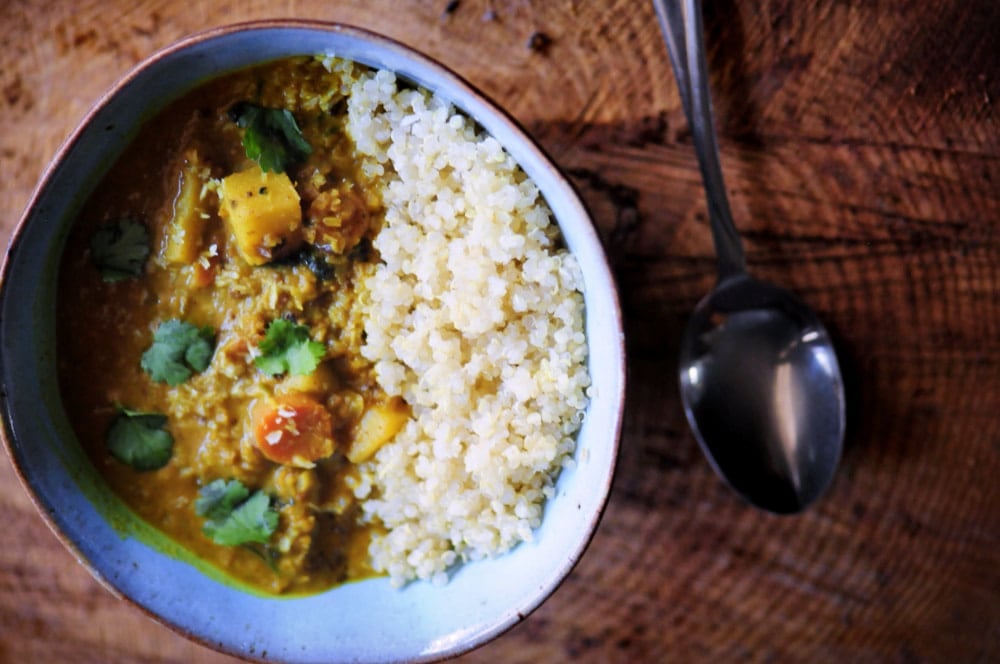 A bowl filled with curry and cooked couscous, garnished with cilantro, placed on a wooden surface next to a metal spoon.