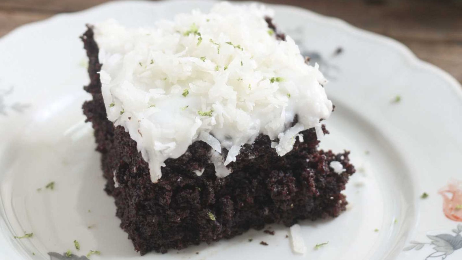 A slice of chocolate cake topped with white coconut shavings on a white floral-patterned plate.