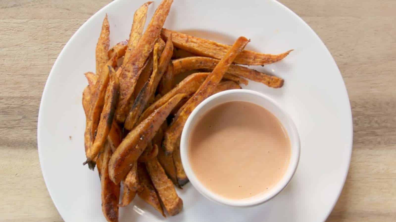 A white plate with a serving of sweet potato fries next to a small cup of dipping sauce, placed on a light wooden surface.