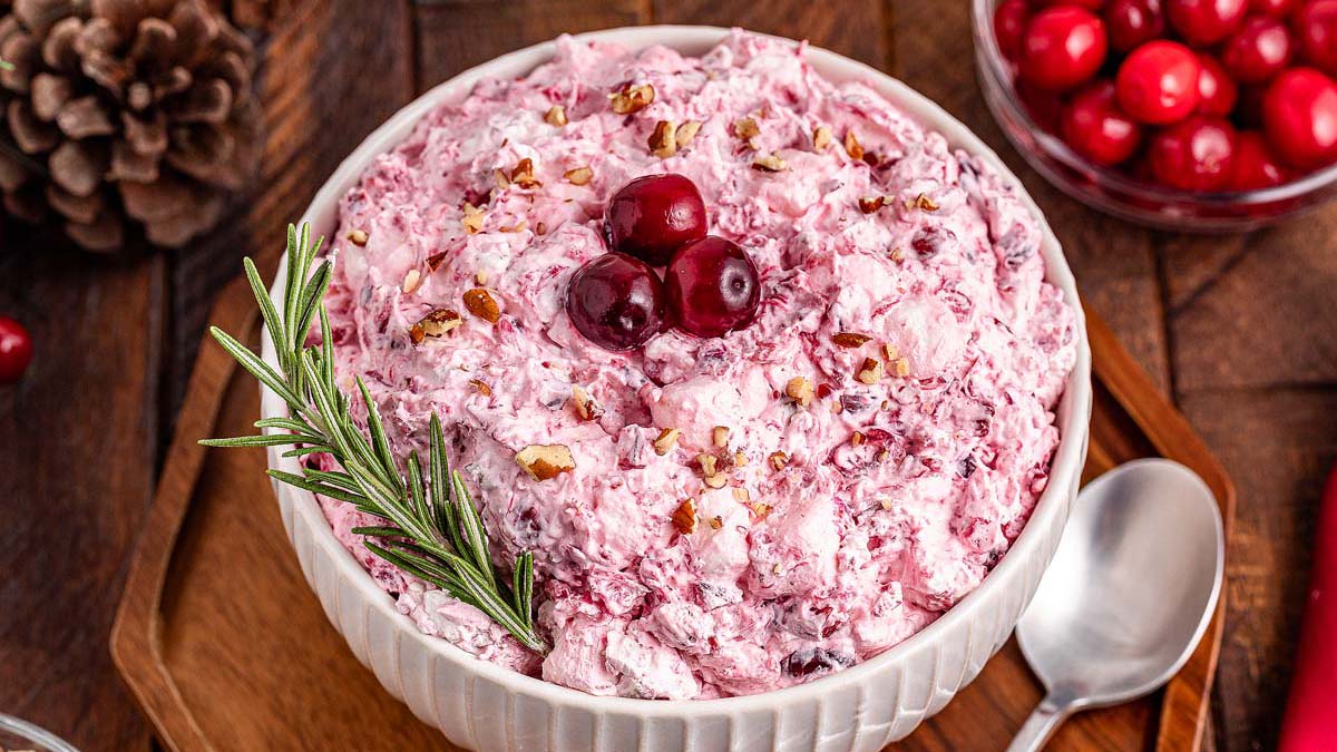 A bowl of pink cranberry fluff salad topped with whole cranberries, chopped nuts, and a sprig of rosemary, with a spoon and cranberries nearby.