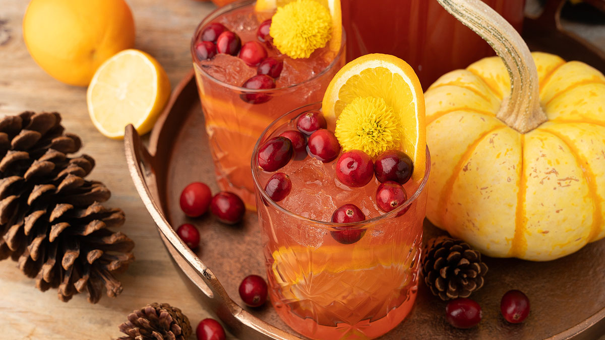 Two glasses of iced cranberry and orange drink are garnished with cranberries, orange slices, and yellow flowers, surrounded by pinecones, cranberries, a pumpkin, and a sliced lemon.