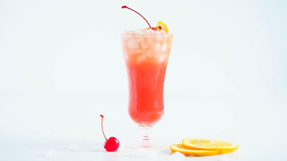 A tall glass filled with a red and orange iced drink, garnished with a cherry and lemon slice, with extra cherry and lemon slices on the white surface nearby.
