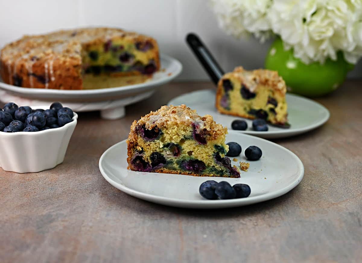 A slice of blueberry coffee cake on a plate with fresh blueberries, a whole cake and another slice in the background, and a white bowl of blueberries nearby.