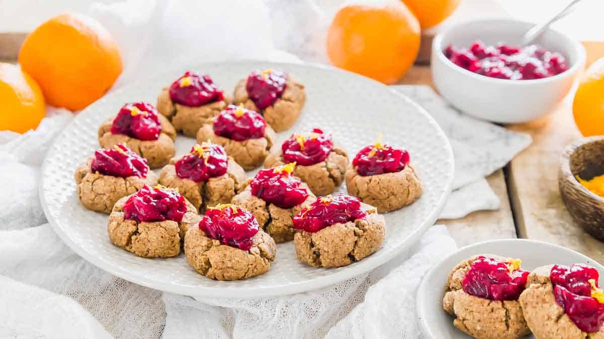 A plate of thumbprint cookies topped with red fruit jam sits on a table, surrounded by oranges and bowls of jam.