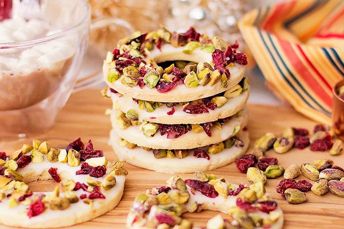 A stack of round shortbread cookies topped with chopped pistachios and dried cranberries sits on a wooden surface.