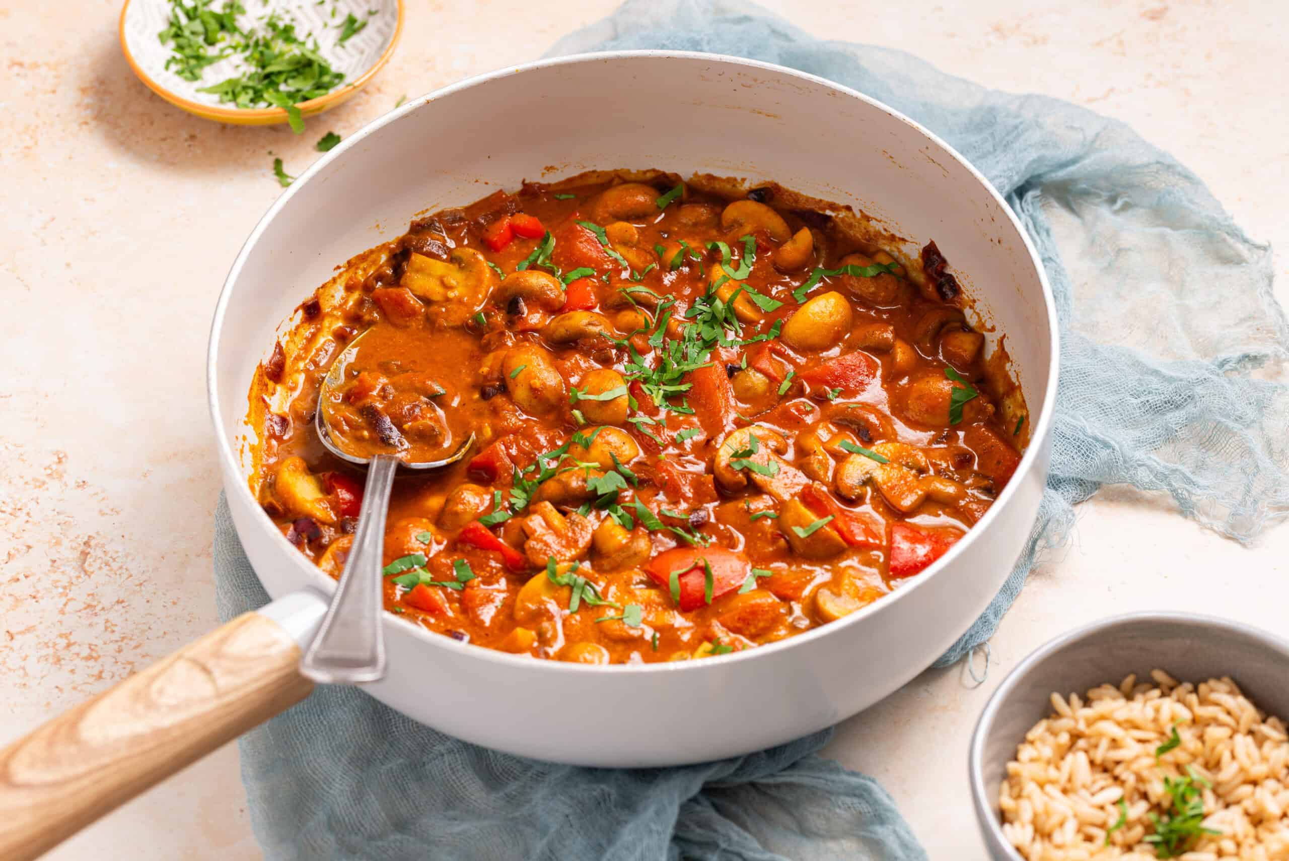 A white pan filled with vegetable curry garnished with chopped herbs, with a metal spoon inside. A bowl of rice and a small dish of herbs are nearby.
