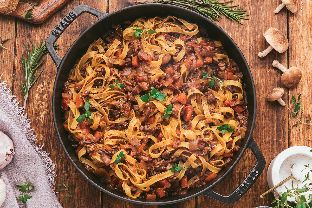 A cast iron pan filled with tagliatelle pasta mixed with a chunky meat and vegetable sauce, garnished with fresh herbs, sits on a wooden table with mushrooms and herbs nearby.
