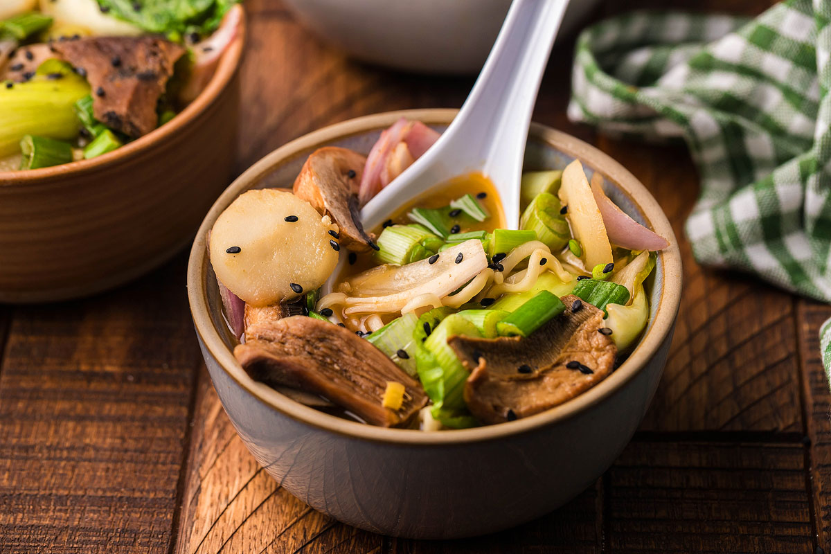 A bowl of noodle soup with mushrooms, greens, sliced vegetables, and black sesame seeds, with a white spoon resting inside.