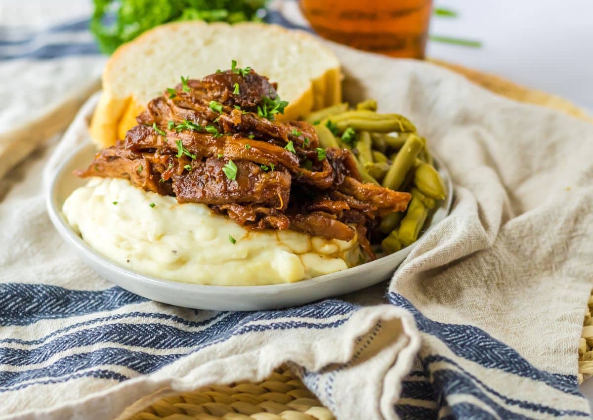 A plate of mashed potatoes topped with shredded beef, served with green beans and slices of bread, placed on a cloth napkin.