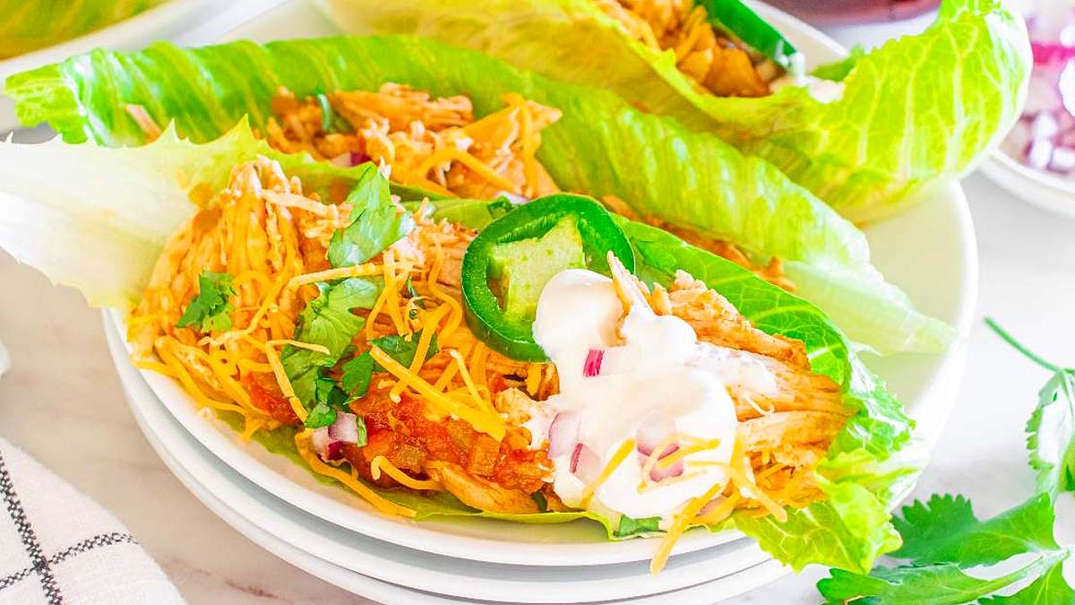 Two lettuce leaves filled with shredded chicken, grated cheese, sliced jalapeรฑo, cilantro, diced red onion, and a dollop of sour cream on a white plate.
