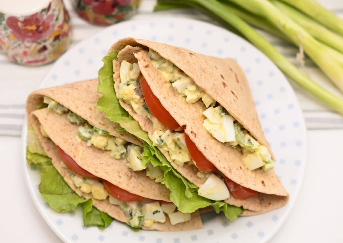 Three folded flatbread wraps filled with lettuce, sliced tomato, and an egg salad mixture on a white plate, with green onions in the background.