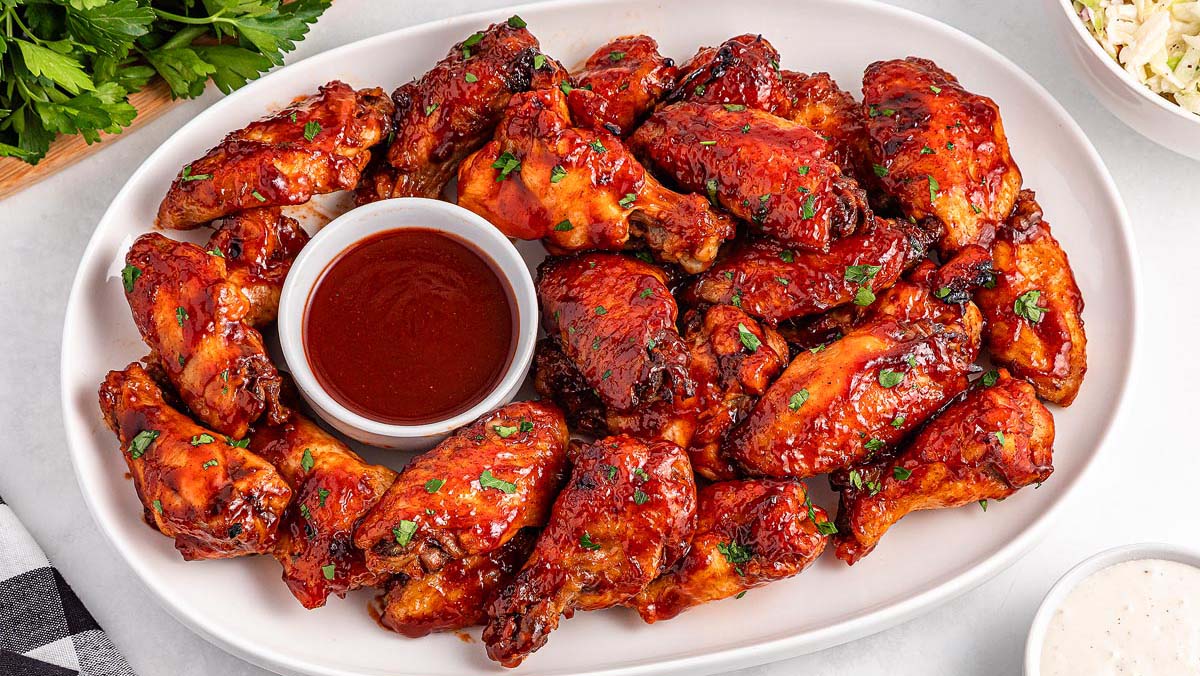 A white platter filled with glazed chicken wings garnished with herbs, surrounding a small bowl of barbecue sauce.