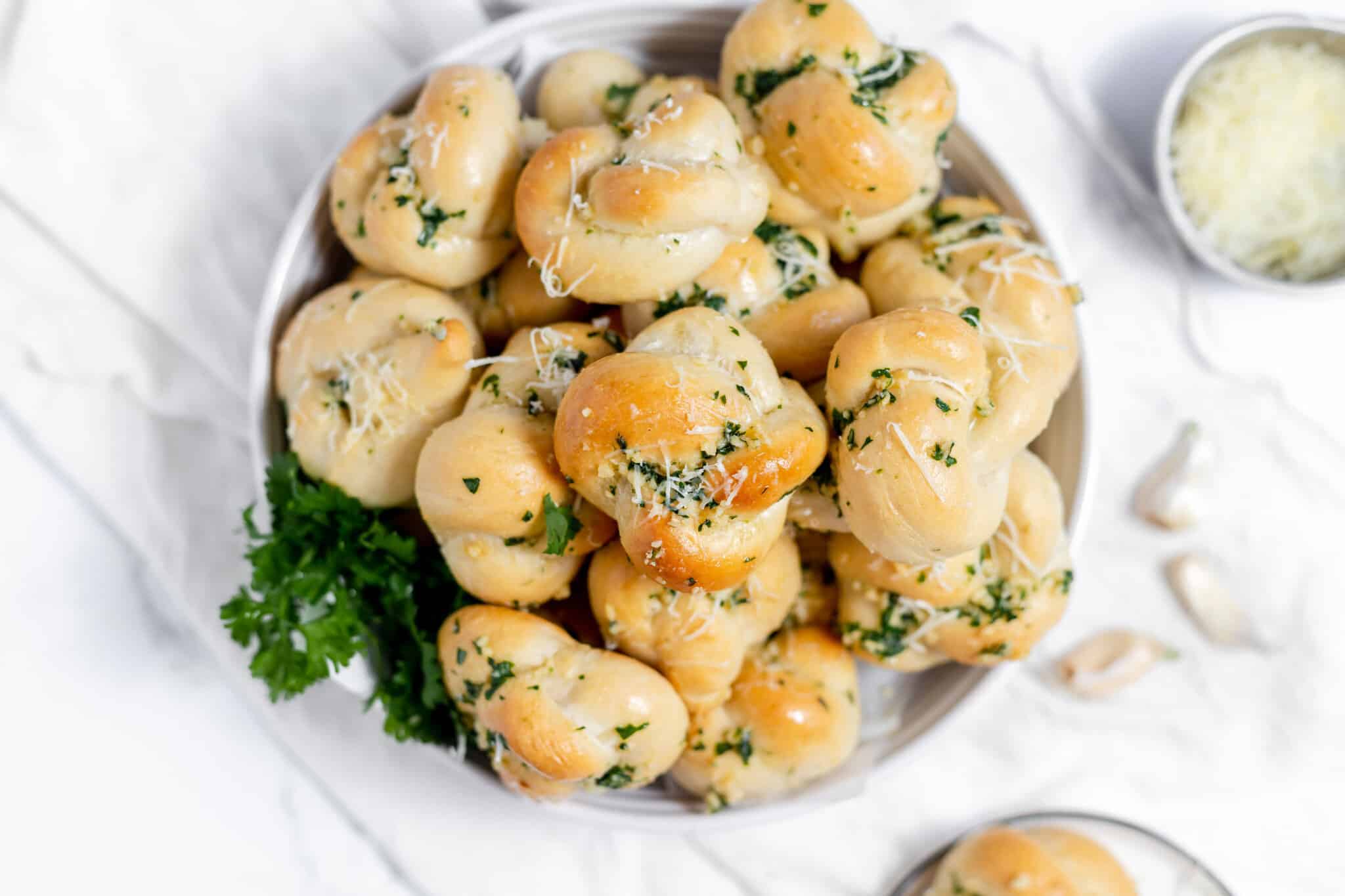 A bowl filled with golden brown garlic knots garnished with chopped parsley and grated cheese, with a small bunch of parsley beside the bowl.
