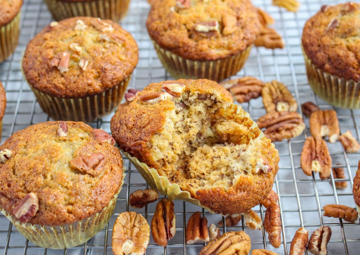Banana nut muffins with chopped pecans sit on a cooling rack; one muffin is broken open to show the moist interior.