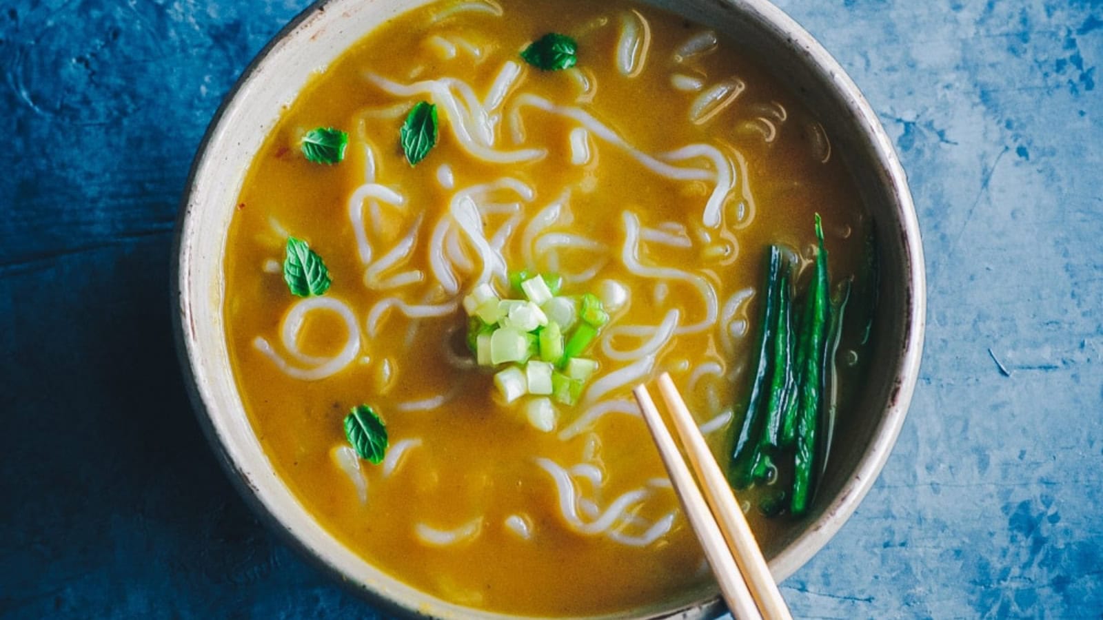 A bowl of noodle soup with green onions, fresh herbs, and green beans, served with chopsticks on a blue surface.