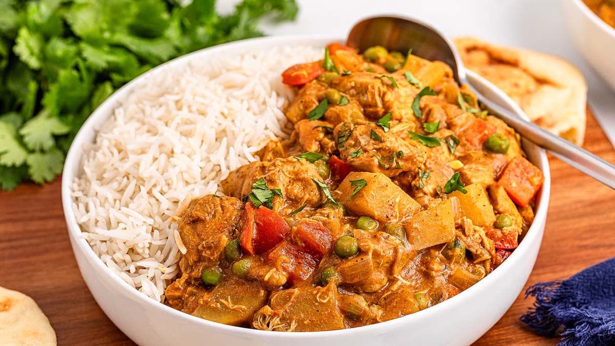 A bowl filled with white rice and vegetable curry, garnished with herbs, with a spoon and naan bread on the side.