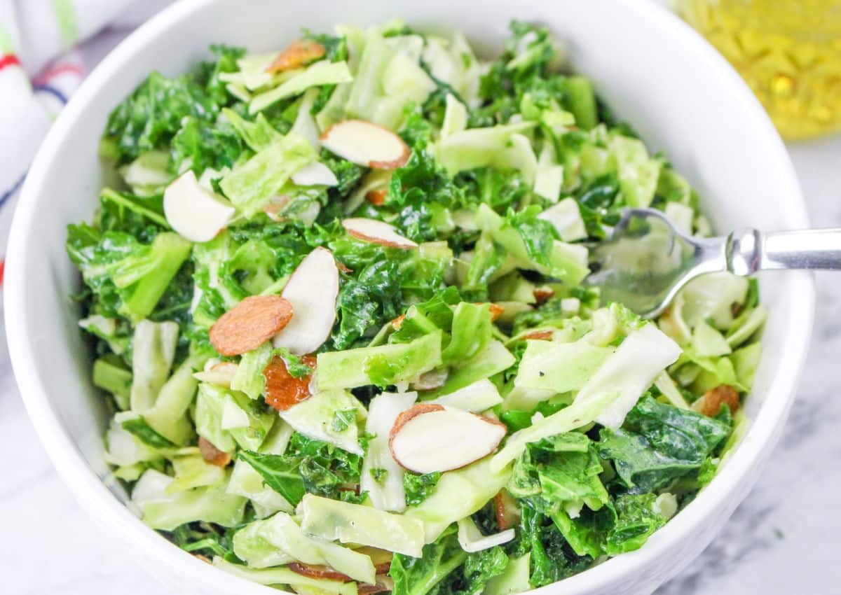 A white bowl filled with a green salad made of chopped leafy vegetables and sliced almonds; a fork rests on the side.
