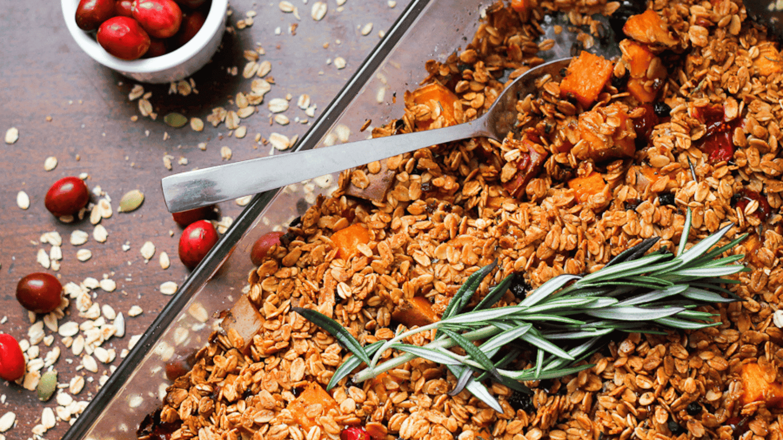 A glass baking dish filled with baked oat and sweet potato crumble, garnished with rosemary, beside a small bowl of cranberries and scattered oats.