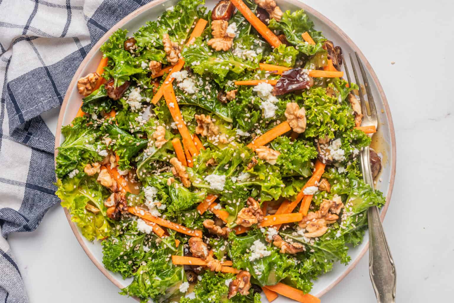 A plate of kale salad with carrot sticks, walnuts, dates, and crumbled cheese, with a fork on the side and a checkered cloth nearby.