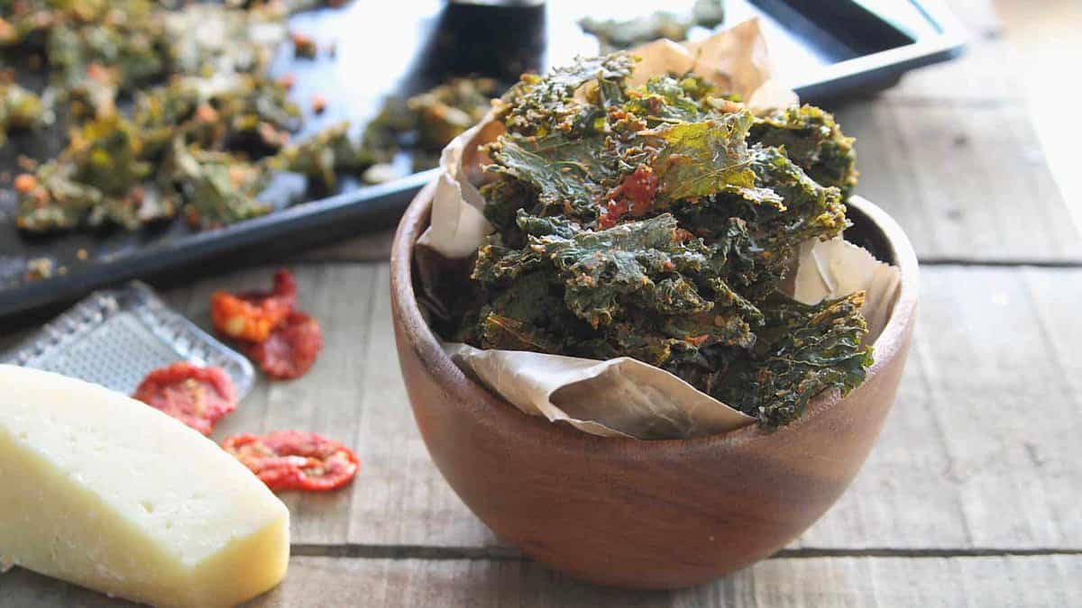 A wooden bowl filled with baked kale chips sits on a wooden table, with cheese, dried tomatoes, and a baking tray of more chips in the background.